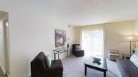 La Fontenay presents a well-appointed single-floor residence designed for comfortable everyday living. The living area serves as the central gathering space, finished with soft wall-to-wall carpeting that adds warmth underfoot. A textured ceiling overhead contributes a classic architectural detail to the room.

Natural light is a defining feature of this space, with a full-height sliding glass door fitted with vertical blinds allowing generous sunlight to filter into the room. The door provides direct access to an outdoor area, creating a seamless connection between interior and exterior living. The neutral wall tones throughout the living area create a versatile backdrop, easily complementing a variety of furnishing styles.

The layout accommodates a functional seating arrangement, with ample room for a sofa, accent chair, and coffee table. A slim console table along one wall offers additional surface space for décor, as illustrated by the colorful floral artwork displayed above it, adding a vibrant focal point to the room. The overall ambiance is light, airy, and inviting, suited to both relaxing and entertaining.