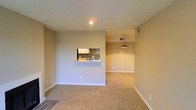 This single-story space is designed with an open-concept layout that promotes a sense of connectivity between the living, dining, and kitchen areas. The living area is finished with wall-to-wall carpet, providing a warm and comfortable foundation underfoot, while tile flooring marks the transition toward the kitchen zone.

A defining architectural feature is the pass-through window cut into the dividing wall between the living area and kitchen. This opening is accented by a granite or stone-look countertop ledge, ideal for casual dining or serving. The kitchen beyond showcases a bold mosaic tile backsplash in dark tones, contrasting with light cabinetry visible through the opening.

The dining area is distinguished by a stylish pendant light that hangs from the ceiling, adding a focal point and warmth to the space. A recessed ceiling light provides additional illumination in the kitchen area. The walls are finished in a neutral beige tone throughout, complemented by clean white baseboards. A textured ceiling adds subtle visual interest. HVAC vents are positioned on the upper walls, ensuring efficient climate control throughout the space.
