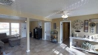 This single-story space presents an open, connected layout highlighted by warm golden-yellow walls and durable tile flooring throughout. The main living area is anchored by a ceiling fan with an integrated light fixture and features a textured popcorn ceiling that adds a classic character to the space. A structural column with a white painted base adds an architectural detail while subtly defining areas within the open floor plan. Natural light filters into the space, casting patterns across the tile floor and creating a bright, welcoming atmosphere. A black metal cabinet provides functional storage and serves as a focal point in the main area, while a decorative wrought-iron étagère holds potted plants, adding a touch of greenery. A large decorative wall clock and a narrow console table with curated décor accessories add personality near one of the doorways. Multiple doorways lead to additional rooms, offering clear sightlines throughout the home. A floor-level vent indicates a central HVAC system, ensuring comfortable climate control. The overall ambiance is casual and livable, with a homey warmth conveyed through the color palette and décor choices.