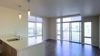 This single-floor residence offers a sleek, contemporary living experience defined by an abundance of natural light and urban views. The open-plan living and dining area is anchored by expansive floor-to-ceiling windows that frame sweeping cityscape panoramas on multiple sides, creating an airy, connected atmosphere. A sliding glass door provides direct access to a private balcony, extending the living space outdoors and offering elevated views of the surrounding cityscape.

The flooring throughout the main living area consists of rich, dark hardwood-style planks that contrast elegantly with the crisp white ceiling and neutral warm-toned walls. A pendant light fixture adds a refined accent above what appears to be a kitchen counter or breakfast bar, which features a light stone or quartz surface and hints at a well-appointed kitchen adjacent to the living space.

Horizontal blinds on the large windows allow for flexible light and privacy control while preserving the clean, modern aesthetic. The overall ambiance is one of understated sophistication — bright, open, and thoughtfully designed, with an emphasis on natural light, outdoor connectivity, and urban living.