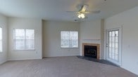 This single-floor residence offers a comfortable and functional layout centered around a spacious living area. The living room serves as the heart of the home, featuring neutral-toned carpeting that extends throughout the space, contributing to a warm and inviting atmosphere. A standout feature is the tiled fireplace with a clean white mantel, providing both a visual focal point and a cozy gathering spot. The room is well-lit by multiple windows, including a centrally positioned window that frames views of mature trees outside, allowing generous amounts of natural light to brighten the interior. A ceiling fan with an integrated light fixture is mounted overhead, offering both air circulation and supplemental lighting. The walls are finished in a soft, neutral tone that complements the overall palette of the space. The layout appears open and unobstructed, allowing for flexible furniture arrangements. A doorway visible to the side of the fireplace suggests easy access to adjacent areas of the home. The overall ambiance is clean, understated, and move-in ready, making it a versatile space suited to a variety of lifestyles.