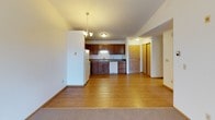 This single-floor unit offers a practical and comfortable layout with a seamless flow between the living and kitchen areas. The open-concept living space features a combination of hardwood-style flooring and plush carpet, creating a natural transition between the kitchen and living room. A chandelier pendant light hangs overhead, adding a touch of warmth to the dining area, while large windows with vertical blinds allow natural light to fill the room. A baseboard heating system runs along the perimeter for efficient climate control.

The kitchen is well-appointed with rich, warm-toned wooden cabinetry both above and below the counter, providing ample storage. It includes a double basin sink, a white dishwasher, electric range, and a full-size refrigerator. A textured ceiling and flush-mount lighting complete the functional space. The entry door is conveniently located adjacent to the kitchen.

The bedroom features soft carpeting, a ceiling fan with light fixture, a window with vertical blinds, and baseboard heating, creating a cozy and comfortable retreat.

The full bathroom includes a shower-tub combination, toilet, and a vanity with a wood cabinet base, single-basin sink, and a lighted mirror above.