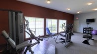 Located at 2335 Atkinson Rd in Biloxi, Mississippi, this single-floor fitness facility offers a dedicated space for exercise and wellness. The workout area is designed with functionality in mind, featuring a mix of cardio and strength training equipment, including an elliptical machine, a cable/weight machine, and additional fitness apparatus arranged to maximize usable floor space. The room is anchored by a row of large windows along one wall, flooding the interior with natural light and providing pleasant views of the surrounding greenery outside. The bold, deep red accent wall adds a energetic and motivating atmosphere to the space, complementing the neutral-toned carpet flooring. Flush-mount ceiling lights provide additional illumination for early morning or evening workouts. The layout allows for comfortable movement between equipment, and the inclusion of a reclining lounge chair suggests a space designed not only for active training but also for cooldown and recovery. Overall, the facility presents a bright, well-equipped environment suited for a variety of fitness routines.