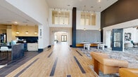 This expansive single-floor amenity space offers a thoughtfully designed layout that blends open gathering areas with distinct, purpose-driven zones. The grand living area serves as the centerpiece, showcasing soaring double-height ceilings, dramatic floor-to-ceiling windows, and an eye-catching mix of warm wood-look and large-format slate tile flooring arranged in a bold striped pattern. Sculptural pendant chandeliers and a stunning tropical palm-leaf wall mural add artistic flair, while abundant natural light pours in through oversized windows framing lush outdoor views.

A billiards table anchors the main gathering space, which flows seamlessly into a stylish bar lounge — branded "Imbibe" — complete with a sleek dark stone island, wine storage, gold pendant lighting, and wood-paneled cabinetry. Adjacent seating areas feature curated contemporary furniture and original artwork throughout.

A more intimate lounge room offers a striking contrast with deep charcoal walls, vibrant green carpet, and moody recessed lighting, creating a cozy, gallery-like atmosphere. The connecting hallway is finished with the same signature flooring and features bold dark doors, including mirrored double barn doors, maintaining design continuity throughout the space.