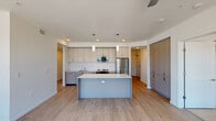 This contemporary single-floor residence is designed with a clean, modern aesthetic and a practical open layout. The kitchen serves as the heart of the home, showcasing flat-front cabinetry in a soft gray-beige tone, a white subway tile backsplash, and stainless steel appliances including a refrigerator and microwave. A spacious central island with a crisp white countertop provides ample prep space and casual seating, while pendant lighting adds a refined touch. Light wood-look flooring extends throughout the kitchen and living areas, contributing to a bright and cohesive atmosphere. The adjacent living area is open and airy, benefiting from recessed lighting and a seamless flow from the kitchen.

The bedrooms are finished with plush neutral carpeting and outfitted with ceiling fans for year-round comfort. Large windows flood the rooms with natural light and frame views of the surrounding landscape. The primary bedroom features dual windows offering expansive outdoor vistas.

Two full bathrooms are appointed with light wood flooring, gray cabinetry with white countertops, and tub-shower combinations. One bathroom includes built-in wall shelving for added storage convenience. The overall palette of whites, grays, and warm wood tones creates a calm, cohesive living environment throughout the space.
