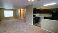 This three-floor home offers a practical and well-organized layout centered around an open-concept main level. The living area is finished with plush neutral carpet and painted in soft green-gray tones, creating a cohesive and understated aesthetic. A ceiling fan is installed overhead, adding both functionality and a casual finishing touch to the space.

The kitchen is positioned adjacent to the living area, separated by a partial wall that maintains an open feel while defining each zone. Dark espresso cabinetry contrasts with light granite-style countertops, and black appliances — including a range, microwave, and dishwasher — provide a uniform, coordinated look. The kitchen transitions to hard surface flooring, offering a practical distinction from the carpeted living space. A peninsula provides additional counter space and casual seating potential.

A hallway visible from the main level leads toward a staircase, connecting the floors and suggesting additional living, sleeping, or utility spaces above and below. The textured ceiling, recessed lighting, and clean white trim throughout contribute to a tidy, move-in-ready presentation across the main level.