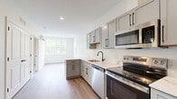 Located at 3 Railroad Street in Newmarket, NH, this thoughtfully designed two-bedroom, one-bathroom unit offers a clean, contemporary aesthetic across a single floor. The kitchen serves as a standout feature, boasting gray shaker-style cabinetry with matte black hardware, light quartz countertops, and a full suite of stainless steel appliances including a range, dishwasher, microwave, and refrigerator. Light hardwood-style flooring runs throughout the main living and kitchen areas, lending warmth and continuity to the space. Natural light filters in through windows, offering views of surrounding greenery. The bedrooms are finished with plush carpeting and neutral wall tones, creating a calm and comfortable atmosphere. The primary bedroom is a well-proportioned retreat, while the second bedroom also benefits from a window with outdoor views. The bathroom is finished with the same cohesive design language — light wood-look flooring, gray cabinetry with black hardware, a white quartz vanity top, and a soaking tub. Built-in baseboard heating and a mini-split system provide year-round climate control. The overall design is modern, clean, and move-in ready, making it an efficient and attractive living space.