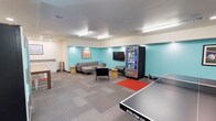 This recreation room is a well-appointed, multi-use space designed to encourage leisure and social interaction. The room is defined by its eye-catching teal accent walls that contrast with the neutral white ceiling and warm beige trim, creating a lively yet comfortable atmosphere. Overhead, recessed and fluorescent lighting ensures the space is well-lit throughout.

At the forefront of the room sits a full-size ping-pong table, serving as a central activity hub for friendly competition. The lounge area features a contemporary gray sectional sofa and additional seating with accent tables, all arranged around a flat-screen television mounted on the teal wall — ideal for unwinding and entertainment. A snack and beverage vending machine is conveniently positioned nearby, adding a practical amenity for guests or residents.

The flooring consists of a patterned carpet with a red area rug anchoring the lounge seating zone, adding warmth and a sense of defined space. A counter or service area is visible along one wall, enhancing the room's functionality. Wall art adds a finishing decorative touch. Overall, this recreation room offers a balanced combination of active entertainment and relaxed lounging in a cheerful, thoughtfully designed environment.