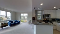 This single-floor space showcases an open-concept layout that integrates the living area and kitchen into one cohesive, functional environment. The living area is finished with plush carpeting in a neutral tone, creating a comfortable and inviting atmosphere. Large double-hung windows along the exterior walls flood the space with abundant natural light while framing pleasant views of the surrounding trees and outdoor scenery.

The kitchen is positioned adjacent to the living area and features a peninsula with a granite countertop, providing both additional workspace and a natural visual divider between the two zones. Cabinetry is finished in white, contributing to the clean, bright aesthetic of the space. Stainless steel appliances, including a microwave and range, add a modern touch, while a built-in sink is integrated neatly into the peninsula countertop.

Recessed lighting is installed throughout the ceiling, supplementing the generous natural light. Crown molding trims the ceiling, adding a refined architectural detail. The overall ambiance is light, airy, and well-suited for both everyday living and entertaining, with a cohesive design language that ties the entire space together.