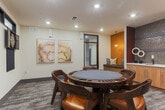 Located at 151 Integra Vistas Dr in Chattanooga, Tennessee, this single-floor game room is thoughtfully designed as a dedicated entertainment and social space. At its center sits a full-size round poker table surrounded by rich dark wood chairs with tan leather upholstered seats, serving as the focal point of the room. The space is anchored by a built-in wet bar niche, framed by a striking textured mosaic tile accent wall in dark metallic tones, complemented by warm amber-hued wall panels on either side. Flanking the wet bar are two built-in bench seating areas with cushioned pads and decorative pillows, ideal for casual conversation or spectating. Recessed ceiling lighting and pendant fixtures cast a warm, inviting glow throughout. On the opposing wall, a wall-mounted flat-screen television and an electronic dartboard with a chalkboard scoreboard provide additional entertainment options. A row of clerestory windows near the ceiling introduces natural light without compromising wall space. The flooring features a neutral-toned patterned carpet that adds texture and warmth underfoot. Abstract artwork decorates the walls, lending a contemporary art-gallery feel. Overall, this game room balances a sophisticated aesthetic with highly functional amenities, making it a premier space for social gatherings and recreational activities.