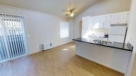 This single-floor space offers a bright and functional open-concept layout that seamlessly connects the living area to the kitchen. The living area features warm wood-look laminate flooring that extends throughout, crown molding along the upper walls, and a ceiling fan with light fixture that enhances air circulation and overhead lighting. A window fitted with blinds allows natural light to fill the room, creating an airy and welcoming atmosphere.

The kitchen is positioned adjacent to the living area and is defined by a peninsula counter with a dark countertop, offering additional workspace and a natural boundary between the two zones. White cabinetry lines the upper and lower kitchen walls, providing ample storage space. The kitchen is equipped with a white range, a built-in microwave above, and a stainless steel sink, all contributing to a clean and classic aesthetic.

The overall color palette is neutral, with white walls and trim that keep the space feeling open and versatile. The layout is efficient and well-organized, making excellent use of the available footprint. The combination of practical finishes, natural light, and an open floor plan makes this a comfortable and functional living space.