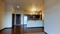 This single-floor residence offers a thoughtfully designed open-concept layout that seamlessly connects the kitchen and living areas. The kitchen is a standout feature, showcasing rich dark cabinetry that extends to the upper walls, complemented by a mosaic tile backsplash and granite countertops. Stainless steel appliances, including a refrigerator, built-in microwave, and range, add a modern touch to the space. A raised breakfast bar with seating overhang provides a casual dining option and serves as a natural divider between the kitchen and the main living area. Four elegant pendant lights hang at varying heights above the kitchen, casting a warm, inviting glow and drawing the eye upward toward the high ceilings. The living area features rich, dark hardwood flooring that runs throughout, adding warmth and continuity to the space. A solid dark-finish entry door with a sidelite is visible from the main living area, grounding the open floor plan. The overall ambiance is modern yet warm, with a neutral color palette on the walls that allows the dark wood tones and stainless steel accents to take center stage, creating a cohesive and inviting interior environment.