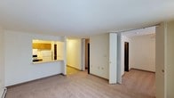 This single-floor space offers a practical, open layout designed for comfortable everyday living. The main living area is finished with wall-to-wall carpeting in a neutral tone, creating a warm and inviting atmosphere. A pass-through counter connects the living space to the kitchen, which features honey-toned wood cabinetry, a white refrigerator, and under-cabinet lighting that adds a soft ambient glow. The kitchen's layout is compact yet functional, making efficient use of available space.

From the main living area, a central hallway provides access to additional rooms, with multiple doorways visible leading to what appear to be bedrooms or utility spaces. Bifold closet doors are a notable feature, offering organized storage solutions along the hallway. The space is finished with consistent white walls and neutral baseboards throughout, contributing to a clean, cohesive look. Overhead lighting keeps the interior well-illuminated. The overall design emphasizes simplicity and functionality, with a straightforward floor plan that connects all areas of the unit in a logical and accessible manner.