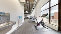 This fitness center offers a comprehensive workout environment thoughtfully organized across a single floor. The space features an industrial-inspired design with exposed concrete ceilings, visible ductwork, and large floor-to-ceiling windows that flood the interior with natural light and provide open views of the surrounding urban environment. The durable rubber flooring throughout provides both safety and functionality for a wide range of exercises.

The main cardio zone is well-appointed with multiple treadmills, stationary cycling bikes, an elliptical, and a stair climber, all positioned to take advantage of the abundant natural light from the expansive windows. A dedicated strength training area features a multi-station power rack, TRX suspension trainers, adjustable benches, and an extensive dumbbell rack, complemented by wall-to-wall mirrors that enhance the sense of space.

A distinct functional training zone is defined by artificial turf flooring, ideal for dynamic movements, agility drills, and ground-based exercises. This area is further equipped with free weights and additional training accessories. A water cooler station is conveniently located within the space to keep users hydrated. The overall ambiance is bright, clean, and modern, creating an energizing atmosphere suitable for a full range of fitness activities.