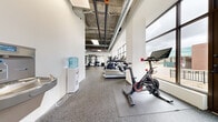 This fitness center offers a comprehensive workout environment thoughtfully organized across a single floor. The space features an industrial-inspired design with exposed concrete ceilings, visible ductwork, and large floor-to-ceiling windows that flood the interior with natural light and provide open views of the surrounding urban environment. The durable rubber flooring throughout provides both safety and functionality for a wide range of exercises.

The main cardio zone is well-appointed with multiple treadmills, stationary cycling bikes, an elliptical, and a stair climber, all positioned to take advantage of the abundant natural light from the expansive windows. A dedicated strength training area features a multi-station power rack, TRX suspension trainers, adjustable benches, and an extensive dumbbell rack, complemented by wall-to-wall mirrors that enhance the sense of space.

A distinct functional training zone is defined by artificial turf flooring, ideal for dynamic movements, agility drills, and ground-based exercises. This area is further equipped with free weights and additional training accessories. A water cooler station is conveniently located within the space to keep users hydrated. The overall ambiance is bright, clean, and modern, creating an energizing atmosphere suitable for a full range of fitness activities.