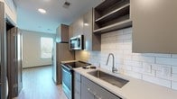 Located at 1005 W Main St in Durham, NC, this single-floor unit offers a thoughtfully designed layout with modern finishes throughout. The galley-style kitchen is a standout feature, equipped with stainless steel appliances including a side-by-side refrigerator with water dispenser, electric range, and built-in microwave. Sleek gray cabinetry with open upper shelving frames a classic white subway tile backsplash, while light quartz countertops and a deep stainless steel sink complete the polished look. Light wood-tone luxury vinyl plank flooring runs consistently throughout the unit, contributing to a cohesive, contemporary aesthetic. The open living area is bright and versatile, with a ceiling fan, neutral wall tones, and an arched pass-through that connects seamlessly to the kitchen space. The bedroom offers a generous layout with access to a closet area. The bathroom is well-appointed with gray vanity cabinetry, a white quartz countertop, an undermount sink, and a large framed mirror. A tiled walk-in shower with a glass door serves as the bathroom's focal point, complemented by wood-look flooring that ties the space together. Natural light filters in through slatted blinds, enhancing the unit's clean, modern ambiance.