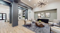This thoughtfully designed single-floor community space combines contemporary aesthetics with functional elegance throughout. Soaring vaulted ceilings and expansive floor-to-ceiling windows flood the interiors with natural light, while light-toned hardwood-style flooring flows seamlessly from room to room, creating a cohesive and welcoming atmosphere.

The main lounge area serves as the visual centerpiece, featuring a striking sputnik chandelier, a geometric wood coffee table, and mid-century modern accent chairs arranged around a patterned area rug. A sage-green accent niche displays botanical artwork above a sleek black credenza, adding a refined, organic touch. Built-in shelving framed in a deep charcoal hue and accented with a marble-surround fireplace wall adds architectural depth and sophistication.

The reception area is anchored by a dark, textured front desk with seating for multiple guests, illuminated by a matching sputnik pendant light and flanked by large windows overlooking an outdoor covered porch and pool area. Integrated display niches with decorative accents enhance the professional yet inviting character of the space.

A separate media and dining area features teal hexagonal tile as a bold accent wall, a wall-mounted flat-screen television, bar-height counter seating, and a dining table surrounded by warm mustard-yellow upholstered chairs. Direct access to the outdoor area is provided through large sliding glass openings, connecting interior and exterior spaces effortlessly.