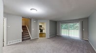 This two-story space is designed with a practical and comfortable layout across both levels. The main floor centers around a generously sized living area finished with plush grey carpeting and neutral grey walls, creating a clean, understated aesthetic. A large sliding glass door floods the space with natural light and provides views of lush greenery outdoors. Adjacent to the living area, the kitchen is well-appointed with white shaker-style cabinetry, granite countertops, stainless steel appliances including a refrigerator, microwave, and range, as well as a dishwasher and hardwood-style flooring. A carpeted staircase leads to the upper floor, where the bedrooms offer a calm retreat. The bedrooms feature the same soft grey carpeting and neutral wall tones, with ample storage provided by built-in bifold closets. A full bathroom on the upper level includes a granite-topped vanity with white cabinetry, a framed mirror with overhead lighting, tile flooring, and a glass-enclosed shower. Throughout both floors, the consistent use of neutral tones, clean finishes, and practical fixtures contributes to a cohesive and functional overall ambiance.