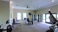 This single-floor fitness room offers a practical and comfortable environment for exercise. The space is outfitted with a selection of strength training equipment, including a multi-station cable machine, adjustable weight benches, and a free-weight setup with plate-loaded barbells, providing options for a full-body workout. Carpeted flooring runs throughout the room, adding warmth underfoot while defining the workout area. Neutral beige walls and crown molding contribute a clean, polished aesthetic. Recessed lighting is complemented by a ceiling fan, ensuring the space remains well-lit and well-ventilated during use. Multiple windows along one wall allow natural light to fill the room and provide views of surrounding greenery, creating an open and airy atmosphere. A wall-mounted flat-screen television is positioned for easy viewing during workouts. A glass door on one side of the room provides additional access and further enhances the sense of openness by allowing natural light to filter in. Overall, the space is thoughtfully designed to balance functionality with a comfortable, inviting environment suitable for regular fitness use.