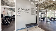 Located at 8130 Prescott Dr in Vienna, Virginia, the Merrifield Fitness Center at Dunn Loring Station offers a spacious, single-floor facility designed to support a comprehensive fitness experience. The center is organized into distinct workout zones that flow seamlessly throughout the open layout. Upon entry, members are greeted by a clean, light-tiled foyer featuring a branded welcome mat and easy access to the main floor.

The strength training area features a robust selection of cable machines, multi-use functional trainers, free weights, barbells, weight plates, and adjustable benches. Diamond-plate steel columns accent the industrial aesthetic, complemented by an exposed ceiling with structural beams, ductwork, and overhead lighting that lend the space a modern, performance-focused atmosphere. Large mirrors line the walls, enhancing the sense of openness.

A dedicated cardio zone houses Life Fitness treadmills and elliptical trainers equipped with individual digital screens. This area benefits from abundant natural light streaming through floor-to-ceiling windows that frame lush outdoor greenery, creating an energizing workout environment with pleasant outdoor views.

The rubber flooring throughout provides both safety and durability, while the bright, neutral color palette keeps the atmosphere clean and professional. Facility policies are clearly posted at the entrance, ensuring a well-managed environment for all members.