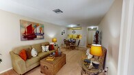 This single-story unit in Kenner, Louisiana, offers a thoughtfully arranged floor plan that maximizes its available space across one bedroom and one bathroom. The layout flows naturally from a carpeted living room through a combined dining area and into a galley-style kitchen, connected by a central hallway that provides access to the bedroom and bathroom.

The living room features neutral-toned walls, carpet flooring, and windows dressed with curtains that allow natural light to filter in. A large display cabinet and open wall space add functional and visual interest to the room.

Adjacent to the living area, the dining space accommodates a round glass-top table and wicker chairs, positioned directly beside the kitchen pass-through. The kitchen is equipped with light wood cabinetry, an electric range, a refrigerator, and wood-style flooring, offering a compact yet complete cooking space with bright yellow accent walls above the counter.

The bedroom features carpet flooring, a ceiling fan, a window with blinds, and ample wall space. The bathroom includes a tub-shower combination with a decorative curtain and wood-style flooring. Throughout the unit, a consistent nautical decorative theme ties each room together cohesively.