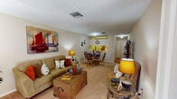 This single-story unit in Kenner, Louisiana, offers a thoughtfully arranged floor plan that maximizes its available space across one bedroom and one bathroom. The layout flows naturally from a carpeted living room through a combined dining area and into a galley-style kitchen, connected by a central hallway that provides access to the bedroom and bathroom.

The living room features neutral-toned walls, carpet flooring, and windows dressed with curtains that allow natural light to filter in. A large display cabinet and open wall space add functional and visual interest to the room.

Adjacent to the living area, the dining space accommodates a round glass-top table and wicker chairs, positioned directly beside the kitchen pass-through. The kitchen is equipped with light wood cabinetry, an electric range, a refrigerator, and wood-style flooring, offering a compact yet complete cooking space with bright yellow accent walls above the counter.

The bedroom features carpet flooring, a ceiling fan, a window with blinds, and ample wall space. The bathroom includes a tub-shower combination with a decorative curtain and wood-style flooring. Throughout the unit, a consistent nautical decorative theme ties each room together cohesively.