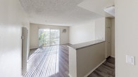 This single-floor unit is designed with an open, functional layout that seamlessly connects the living area and kitchen. The living space features wood-look hardwood-style flooring and neutral-toned walls, creating a bright and clean atmosphere. A textured popcorn ceiling runs throughout, adding a classic touch to the interior. A large sliding glass door with vertical blinds allows abundant natural light to fill the living area while providing outdoor access and views of surrounding greenery. A wall-mounted air conditioning unit serves the living space.

The kitchen is defined by a peninsula counter with a granite-style surface, offering both workspace and a natural divider between the kitchen and living areas. Cream-colored upper cabinetry, a dark tile backsplash, and a white gas range contribute to a practical and tidy cooking environment. A microwave is integrated above the counter space.

The bathroom includes a vanity with a granite-style countertop, a toilet, a bathtub with shower fixtures, and a chrome towel bar mounted on the wall, maintaining the unit's consistent neutral color palette. Overall, the space offers a straightforward, well-lit, and practical living environment.