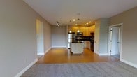 This single-floor residence offers a thoughtfully designed open-concept layout that seamlessly blends the living and kitchen areas into one cohesive space. The living area features plush carpeting that transitions smoothly into warm hardwood-style flooring extending through the rest of the space, creating a welcoming and comfortable atmosphere.

The kitchen is a standout feature, showcasing light wood cabinetry that extends to the ceiling, complemented by dark granite countertops and a matching kitchen island with a contrasting white base. A decorative tile backsplash adds visual interest, while stainless steel appliances — including a refrigerator, range, and dishwasher — provide a sleek, modern finish. Pendant lighting hangs above the island, adding both style and functionality, and recessed lighting throughout ensures the space is well-lit.

The open layout allows for a natural flow between the kitchen and living areas, making the space ideal for entertaining or everyday living. Neutral gray walls provide a clean, versatile backdrop throughout, while track lighting in the living area adds a modern, flexible lighting solution. White baseboards and trim offer a crisp, polished finish. Overall, the space combines contemporary design elements with practical functionality for comfortable, stylish living.