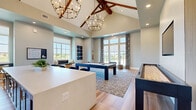 Located at 13000 Saginaw Blvd in Fort Worth, Texas, this game room is a well-appointed recreational space designed for socializing and entertainment. The room is defined by its dramatic vaulted ceiling, accented with rich, exposed wood beam trusses that add a warm, rustic contrast to the otherwise modern interior. Two statement chandelier light fixtures hang from the peak, casting an inviting glow throughout the space.

The open floor plan centers around a navy blue pool table set atop a patterned area rug, with a full-length shuffleboard table positioned alongside it. Light wood-look tile flooring extends across the room, complementing the neutral, soft gray wall tones. A built-in wet bar with a white countertop, bar seating, and wall-mounted flat-screen television provides a dedicated lounge area ideal for gathering.

Large floor-to-ceiling windows and French doors line the walls, flooding the interior with natural light and offering open views of the surrounding landscape. A cozy built-in window bench with cushions creates a comfortable nook for relaxing. Decorative wall art and a wall-mounted cue rack add thoughtful finishing touches. The overall ambiance is modern yet welcoming, blending recreational functionality with refined design throughout.