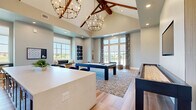 Located at 13000 Saginaw Blvd in Fort Worth, Texas, this game room is a well-appointed recreational space designed for socializing and entertainment. The room is defined by its dramatic vaulted ceiling, accented with rich, exposed wood beam trusses that add a warm, rustic contrast to the otherwise modern interior. Two statement chandelier light fixtures hang from the peak, casting an inviting glow throughout the space.

The open floor plan centers around a navy blue pool table set atop a patterned area rug, with a full-length shuffleboard table positioned alongside it. Light wood-look tile flooring extends across the room, complementing the neutral, soft gray wall tones. A built-in wet bar with a white countertop, bar seating, and wall-mounted flat-screen television provides a dedicated lounge area ideal for gathering.

Large floor-to-ceiling windows and French doors line the walls, flooding the interior with natural light and offering open views of the surrounding landscape. A cozy built-in window bench with cushions creates a comfortable nook for relaxing. Decorative wall art and a wall-mounted cue rack add thoughtful finishing touches. The overall ambiance is modern yet welcoming, blending recreational functionality with refined design throughout.