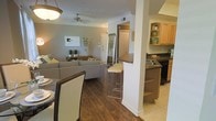 This single-story residence showcases an open-concept layout that integrates the dining, living, and kitchen areas into one cohesive, flowing space. Rich hardwood-style flooring runs throughout the main living and dining areas, transitioning to light-toned tile in the kitchen zone, creating a natural visual distinction between spaces.

The living area is generously proportioned, featuring neutral-toned walls, crown molding details, and a ceiling fan that contributes to both comfort and style. A structural column with a breakfast bar anchors the transition between the living space and kitchen, offering counter-height seating for casual dining or entertaining.

The kitchen is well-appointed with light wood cabinetry featuring open display shelving, dark countertops, a sink, and black appliances. The contrast between the cabinetry and countertops adds visual depth to the space.

The dining area, positioned near the entry, accommodates a full dining set and benefits from the open sightlines throughout the home. The entry features a full-length mirror and access to additional areas of the home. Overall, the space presents a warm, functional, and well-connected interior ideal for everyday living.