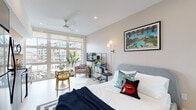 This thoughtfully designed studio unit offers an open-concept layout that seamlessly integrates the living, sleeping, and dining areas into one cohesive space. Large floor-to-ceiling windows flood the interior with natural light, offering views of surrounding urban architecture, while roller shades provide privacy as needed. A ceiling fan overhead ensures year-round comfort.

The living area is furnished with a cozy seating arrangement and an open shelving unit that doubles as an entertainment and display space. Adjacent to it, a compact dining area features a round marble-top table with vibrant accent chairs, adding a pop of color to the neutral palette. The sleeping area is anchored by a plush upholstered bed with eclectic decorative pillows, complemented by globe wall sconces and curated wall art. A walk-in closet with built-in shelving and hanging storage provides ample organization.

The kitchen is outfitted with two-tone cabinetry — light wood upper cabinets paired with gray lower units — dark countertops, a textured tile backsplash, and a full suite of stainless steel appliances. The bathroom features large-format gray stone tiles, a wall-mounted floating vanity, and clean white walls, creating a spa-like atmosphere. Light hardwood flooring in the entryway transitions smoothly into the main living area, tying the entire unit together with warmth and continuity.