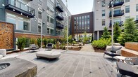 Located at 7970 Maitland St in McLean, Virginia, this thoughtfully designed outdoor amenity space serves as a central gathering hub within a modern mixed-use community. The space is organized across several distinct zones, each offering a unique environment for relaxation and recreation.

A spacious covered terrace features wood-look tile flooring and houses a Kettler ping-pong table alongside colorful picnic-style dining furniture, all open to panoramic views of the surrounding development. An adjacent semi-enclosed lounge area includes a built-in linear fireplace, wall-mounted television, and warm wood accent walls paired with contemporary gray upholstered seating.

The central courtyard is a landscaped sanctuary, featuring large-format slate-toned pavers, curved lounge seating, and a striking natural stone fire pit. String lights overhead and a decorative laser-cut metal screen panel add architectural character and ambiance after dark.

Additional garden areas incorporate mature plantings, natural boulders, and specimen trees set alongside floor-to-ceiling glass curtain walls. A separate patio zone offers umbrella-shaded seating on broad stone pavers, ideal for outdoor gatherings. The cohesive design blends natural materials, contemporary architecture, and varied seating environments throughout.