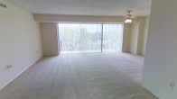 This single-floor space offers a generously sized living area defined by its clean, minimalist design and an abundance of natural light. The most striking feature is the expansive wall of floor-to-ceiling windows fitted with vertical blinds, which flood the interior with daylight while framing lush greenery and outdoor views beyond a railed balcony. The seamless connection between the interior and the outdoors creates an airy, open atmosphere throughout the space.

The living area is finished with wall-to-wall carpeting in a neutral beige tone, lending warmth and a soft underfoot feel that complements the light-colored walls. A ceiling fan is positioned overhead, providing practical air circulation and subtle overhead lighting to enhance comfort throughout the seasons.

The overall aesthetic is understated and versatile, with a neutral color palette that allows for a wide range of furnishing styles. The open layout provides ample room for flexible furniture arrangements, making the space well-suited for both relaxed everyday living and entertaining. The combination of natural light, outdoor connectivity, and open design gives the space a welcoming and comfortable ambiance.