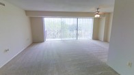 This single-floor space offers a generously sized living area defined by its clean, minimalist design and an abundance of natural light. The most striking feature is the expansive wall of floor-to-ceiling windows fitted with vertical blinds, which flood the interior with daylight while framing lush greenery and outdoor views beyond a railed balcony. The seamless connection between the interior and the outdoors creates an airy, open atmosphere throughout the space.

The living area is finished with wall-to-wall carpeting in a neutral beige tone, lending warmth and a soft underfoot feel that complements the light-colored walls. A ceiling fan is positioned overhead, providing practical air circulation and subtle overhead lighting to enhance comfort throughout the seasons.

The overall aesthetic is understated and versatile, with a neutral color palette that allows for a wide range of furnishing styles. The open layout provides ample room for flexible furniture arrangements, making the space well-suited for both relaxed everyday living and entertaining. The combination of natural light, outdoor connectivity, and open design gives the space a welcoming and comfortable ambiance.