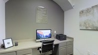 This single-floor business center is a purpose-built workspace designed to meet essential professional and productivity needs. The room features a built-in, countertop-style desk that spans the width of the workspace, providing ample surface area for work tasks. The workstation is equipped with an iMac desktop computer and a laser printer, offering convenient access to computing and printing resources. Beneath the counter, integrated cabinetry provides practical storage solutions, keeping the space organized and clutter-free. The room's neutral grey walls create a calm, focused atmosphere, while an elegant arched architectural detail above the desk adds a refined, polished touch to the functional space. A framed "Business Center Rules" placard is displayed prominently on the wall, guiding users on proper etiquette and usage policies. A comfortable, ergonomic office chair with a padded back and chrome frame ensures a comfortable working experience. The overall design is clean, minimal, and efficient, making this business center a practical and accessible resource for those seeking a professional environment for their work-related tasks.