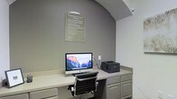 This single-floor business center is a purpose-built workspace designed to meet essential professional and productivity needs. The room features a built-in, countertop-style desk that spans the width of the workspace, providing ample surface area for work tasks. The workstation is equipped with an iMac desktop computer and a laser printer, offering convenient access to computing and printing resources. Beneath the counter, integrated cabinetry provides practical storage solutions, keeping the space organized and clutter-free. The room's neutral grey walls create a calm, focused atmosphere, while an elegant arched architectural detail above the desk adds a refined, polished touch to the functional space. A framed "Business Center Rules" placard is displayed prominently on the wall, guiding users on proper etiquette and usage policies. A comfortable, ergonomic office chair with a padded back and chrome frame ensures a comfortable working experience. The overall design is clean, minimal, and efficient, making this business center a practical and accessible resource for those seeking a professional environment for their work-related tasks.