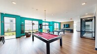Located at 110 Woodcrest Rd in Cherry Hill, NJ, this contemporary amenity space delivers a dynamic and well-appointed social environment across a single, generously sized floor. The layout flows across two distinct living areas, each thoughtfully designed to serve different recreational and social functions.

The game room is a vibrant centerpiece, featuring bold teal accent walls, warm hardwood-style flooring, and an abundance of natural light pouring in through large windows with outdoor views. It is equipped with a pool table, air hockey table, pinball machine, and an arcade game, complemented by a wall-mounted TV and lounge seating. Overhead, a sleek geometric pendant light anchors the space.

The adjacent social lounge area offers a more relaxed atmosphere, furnished with tufted leather sofas and armchairs arranged around a white marble coffee table atop a teal area rug. A slatted wood accent wall frames a fireplace and large TV, adding warmth and texture to the design. A striking multi-arm brass and black chandelier serves as a focal point overhead.

A glass-walled conference and bar-height seating area divides the zones, offering views into a dramatic atrium below. Additional highlights include elevator access, a large-format Scrabble board, and an overall open, airy ambiance defined by clean lines, modern fixtures, and vibrant color accents throughout.