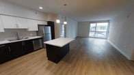 This single-floor space showcases a contemporary open-concept layout that seamlessly connects the kitchen and living areas. The kitchen is outfitted with dark cabinetry contrasted by a bright white countertop island, stainless steel appliances including a refrigerator and dishwasher, and pendant lighting that hangs above the island to create a focused, functional workspace. White upper cabinets provide additional storage while maintaining a clean, modern aesthetic.

The kitchen flows effortlessly into the expansive living area, which is finished with warm wood-look luxury vinyl plank flooring throughout. Large floor-to-ceiling windows along the far wall of the living space flood the interior with abundant natural light and offer views to the exterior. The open layout creates a generous, airy feel that makes the space well-suited for both everyday living and entertaining. Neutral wall tones complement the modern finishes throughout, contributing to a bright and cohesive atmosphere. A secondary entrance or hallway is visible in the background, suggesting additional rooms beyond the main living area.