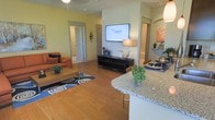 This single-story residence features an open-concept layout that connects the kitchen, dining, and living areas into one cohesive, functional space. The kitchen is anchored by a granite-topped breakfast bar with seating, providing both a casual dining option and a natural divider between the cooking and living zones. Warm hardwood flooring runs throughout, lending continuity and a welcoming tone to the entire main area.

The living room is generously proportioned, accommodating a full seating arrangement centered around a wall-mounted flat-screen television positioned above a sleek media console. Soft yellow-toned walls contribute to the bright, airy atmosphere, while recessed lighting and ceiling fans support both comfort and illumination. A bold patterned area rug adds a decorative focal point to the seating area.

Multiple doorways visible from the main living space indicate access to additional rooms, suggesting a well-organized floor plan with clear separation between the communal and private areas. An arched hallway on one side adds an architectural detail that breaks the linearity of the space. Overall, the layout balances openness with defined functional zones, making it well-suited for both everyday living and entertaining.