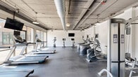 Located at 23511 Chagrin Blvd in Beachwood, Ohio, this fitness center offers a spacious, single-floor exercise facility designed to support a comprehensive workout experience. The space features an industrial-chic aesthetic, with exposed concrete ceilings, large silver ductwork, and pendant lighting that lend the room a modern, loft-like character.

The layout is thoughtfully organized into distinct workout zones. A dedicated cardio area houses treadmills, elliptical machines, and stationary bikes, while a separate cycling studio with floor-to-ceiling glass walls floods the space with natural light and outdoor views. The strength training section features a full rack of dumbbells displayed on tiered racks alongside a large wall mirror, benches, a cable machine, and a power rack with weight plates and medicine balls.

Rubber flooring throughout provides both durability and comfort underfoot. Mounted flat-screen televisions are positioned throughout the space to keep users engaged during workouts. Structural columns are integrated cleanly into the design, defining zones without disrupting the open feel. The generous ceiling height and large windows enhance the sense of openness and airiness across the entire facility, making it an inviting environment for fitness of all kinds.