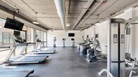 Located at 23511 Chagrin Blvd in Beachwood, Ohio, this fitness center offers a spacious, single-floor exercise facility designed to support a comprehensive workout experience. The space features an industrial-chic aesthetic, with exposed concrete ceilings, large silver ductwork, and pendant lighting that lend the room a modern, loft-like character.

The layout is thoughtfully organized into distinct workout zones. A dedicated cardio area houses treadmills, elliptical machines, and stationary bikes, while a separate cycling studio with floor-to-ceiling glass walls floods the space with natural light and outdoor views. The strength training section features a full rack of dumbbells displayed on tiered racks alongside a large wall mirror, benches, a cable machine, and a power rack with weight plates and medicine balls.

Rubber flooring throughout provides both durability and comfort underfoot. Mounted flat-screen televisions are positioned throughout the space to keep users engaged during workouts. Structural columns are integrated cleanly into the design, defining zones without disrupting the open feel. The generous ceiling height and large windows enhance the sense of openness and airiness across the entire facility, making it an inviting environment for fitness of all kinds.