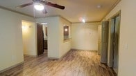 This single-floor residence offers a practical and functional layout designed for comfortable everyday living. The main living area features light wood-style flooring that extends throughout, creating a warm and cohesive feel across the space. A ceiling fan with integrated lighting is centrally mounted, providing both air circulation and ambient illumination.

The living area flows openly toward the kitchen, which is accessible via a structural column-framed opening. A kitchen pass-through with a raised breakfast bar counter connects the two spaces, allowing for convenient interaction between the kitchen and living areas. The kitchen is equipped with dark wood cabinetry and a pendant light fixture, adding a subtle contrast to the light-toned walls and flooring.

The walls throughout are finished in a soft, neutral cream tone, complemented by clean white baseboards and crown molding that lend a polished appearance. Additional doorways visible from the main area suggest access to further rooms, including what appears to be a utility or storage area with built-in shelving. Recessed and track lighting supplements the natural ceiling fixtures, ensuring the space is well-lit. Overall, the layout is straightforward and efficiently organized, making it easy to navigate and utilize.