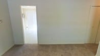 This single-floor space presents a straightforward and functional layout with a calm, neutral aesthetic throughout. The interior is finished with light-toned walls that create a bright, open feel, complemented by wall-to-wall carpeting in a warm beige tone that adds a sense of comfort underfoot. An open doorway connects the main carpeted room to an adjoining area, which features a lighter flooring surface and is illuminated by natural light streaming through large vertical blinds. The vertical blinds suggest access to an outdoor-facing window or sliding door, allowing ample daylight to filter into the space. The overall ambiance is clean, minimal, and move-in ready, with a neutral color palette that provides a versatile backdrop for a variety of furnishing styles. The connected layout between rooms promotes an easy flow throughout the unit, while the simple architectural finishes keep the space feeling uncluttered and practical. This space is well-suited for those seeking a no-frills, functional environment with the foundational elements already in place.