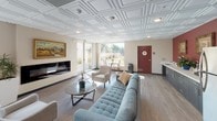 Located in Los Angeles, CA, this single-floor community space is thoughtfully designed to foster comfort and social interaction. The open-plan layout seamlessly connects multiple functional zones, creating a sense of spaciousness throughout.

The lounge area serves as the heart of the space, anchored by a tufted blue sofa, a glass-topped coffee table, and a wall-mounted flat-screen television. Elegant coffered tile ceilings add architectural character overhead, while light wood-look flooring runs throughout, lending a clean and cohesive aesthetic. Large sliding glass doors and floor-to-ceiling windows flood the interior with natural light and offer views of the surrounding outdoor greenery.

Multiple dining areas provide flexible seating arrangements, including a notable solid stone-top dining table surrounded by wooden chairs, ideal for group gatherings. A warm accent wall in deep burgundy adds a touch of sophistication, complemented by framed artwork and decorative open shelving units displaying curated décor pieces.

The kitchen area features stainless steel appliances, including a full-size refrigerator, and provides ample counter space. Subtle touches of greenery throughout the space, including potted plants, enhance the welcoming atmosphere. Restroom facilities are also accessible within the space, and an outdoor patio area is visible through the sliding doors, extending the usable space outdoors.