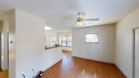 This single-floor space is designed with an open, functional layout that connects the main living and dining areas seamlessly. Warm wood-look flooring runs throughout the unit, complementing the crisp white walls and creating a cohesive, inviting atmosphere. Ceiling fans with integrated lighting fixtures are installed in the primary living areas, providing both comfort and illumination.

The living area is generously proportioned, bathed in natural light from multiple windows fitted with blinds, and features a sliding glass door that opens to an outdoor area. A half-wall partition with a dark granite-style countertop separates the kitchen from the living space, maintaining an open feel while defining each zone.

The kitchen is well-appointed with warm wood cabinetry, dark granite-look countertops, a white subway tile backsplash, and black appliances including an electric range, over-the-range microwave, and refrigerator.

A hallway leads to the bedroom, which features two sets of double-door closets offering ample storage, along with a window that draws in natural light. The bathroom includes a wood-tone vanity with a wide mirror, a tile-style floor, and a clean, functional layout.