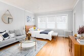 This single-floor space offers a thoughtfully designed layout that combines classic architectural character with mid-century modern furnishings. The main living and sleeping area is generously proportioned, featuring warm hardwood floors, crown molding, and a row of wide windows that flood the room with natural light and offer views of outdoor greenery. A cast-iron radiator underscores the building's classic heritage, while the soft grey walls and carefully curated décor — including a walnut credenza, glass-topped coffee table, and coordinated artwork — create a cohesive and inviting atmosphere. A seating area with a grey upholstered sofa flows naturally alongside the sleeping quarters, making efficient use of the open layout. The kitchen area is equipped with a stainless steel refrigerator, a utility sink with granite-look countertops, and white cabinetry with open shelving — providing functional storage and prep space. The light marble-patterned tile flooring in the kitchen contrasts with the warm hardwood in the living area. Throughout the space, attention to detail is evident in the clean white trim, neutral color palette, and well-placed greenery, resulting in a functional yet stylish environment with a calm, residential ambiance.