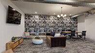 This thoughtfully designed amenity space combines contemporary aesthetics with dynamic, personality-driven décor across a well-appointed single floor. The open-plan interior unfolds across several distinct zones, each with its own character yet unified by patterned carpet flooring and crisp white walls throughout.

A standout lounge area features an eye-catching floor-to-ceiling boombox-print accent wall, exposed metallic ductwork, and a mix of casual seating including a rich leather sofa, woven chairs, and a sculptural round ottoman — creating an inviting, urban-inspired retreat. Nearby, a dramatic floor-to-ceiling dark tile fireplace wall serves as a striking focal point, flanked by vibrant yellow lounge chairs and a round wood coffee table.

A lush, full-height living green wall introduces a refreshing natural element, complementing the bright and airy atmosphere enhanced by skylights and modern globe pendant chandeliers. A flexible co-working area provides long communal tables with seating, ideal for productive gatherings.

The entry area features elegant botanical wallpaper and sleek tile flooring, while a media nook offers a wall-mounted television and casual seating. Outdoors, two distinct spaces extend the amenities further — a spacious courtyard patio with ample lounge seating beside a mixed-material brick wall, and an elevated terrace offering open views of the surrounding neighborhood and tree canopy beyond.