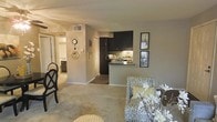 This single-floor residence offers a thoughtfully designed open-concept layout that seamlessly connects the living area to the kitchen. The living space features wall-to-wall carpeting in a neutral tone, creating a warm and comfortable atmosphere. Decorative touches, including framed artwork and a classic wall clock, add character to the space.

The kitchen is a standout feature, showcasing dark espresso-toned cabinetry that contrasts elegantly with a decorative tile backsplash. A peninsula-style breakfast bar provides both additional counter space and a casual dining or gathering spot, making it a functional and inviting area. Under-cabinet lighting enhances visibility and adds a subtle ambiance to the kitchen.

A hallway visible from the living area leads to additional rooms, suggesting a practical and well-organized floor plan. The neutral gray walls throughout provide a versatile backdrop that complements various décor styles. The space benefits from a centralized HVAC system with ceiling vents, ensuring year-round comfort. Overall, the interior presents a cohesive, livable environment with a balance of functionality and style.