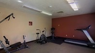 This single-level fitness room offers a dedicated space for exercise and physical activity. The room features a neutral beige color scheme complemented by a bold terracotta-red accent wall, creating an energizing atmosphere suited to a workout environment. Flooring consists of low-pile carpet with a darker rubber mat section beneath the equipment, providing stability and impact absorption during use. The textured ceiling is equipped with fluorescent lighting fixtures that ensure adequate illumination throughout the space, along with a central HVAC vent for climate control. The room is outfitted with several pieces of cardio and resistance equipment, including an air resistance fan bike, a treadmill by TRUE Fitness, and a freestanding resistance/cable machine. A wall-mounted informational sign near the entrance provides facility rules or guidelines for users. The layout allows for comfortable movement between equipment. Overall, the space presents a straightforward and functional fitness environment, well-suited for cardio workouts and general conditioning within a shared or community facility setting.